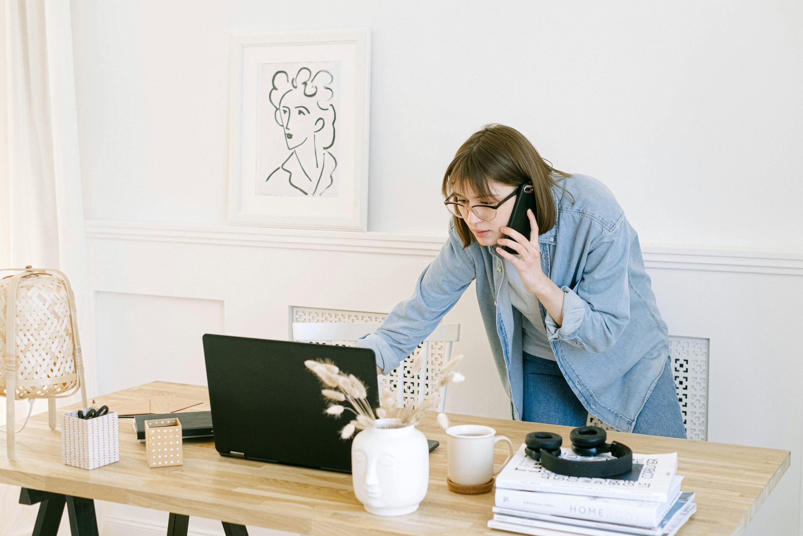 Woman multitasking with laptop and phone in modern home office setting.