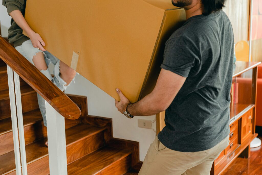 A couple carrying a large cardboard box up the stairs in their new home.