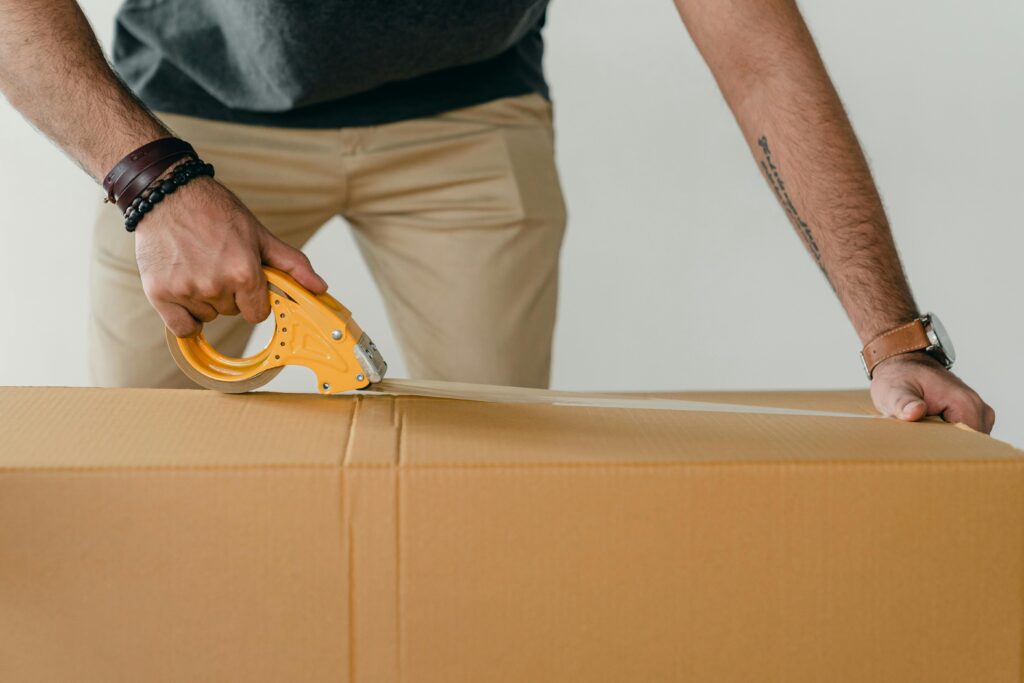pexels-photo-4246118-4246118-1 Crop anonymous young male packer in casual clothes using tape gun dispenser for sealing cardboard boxes