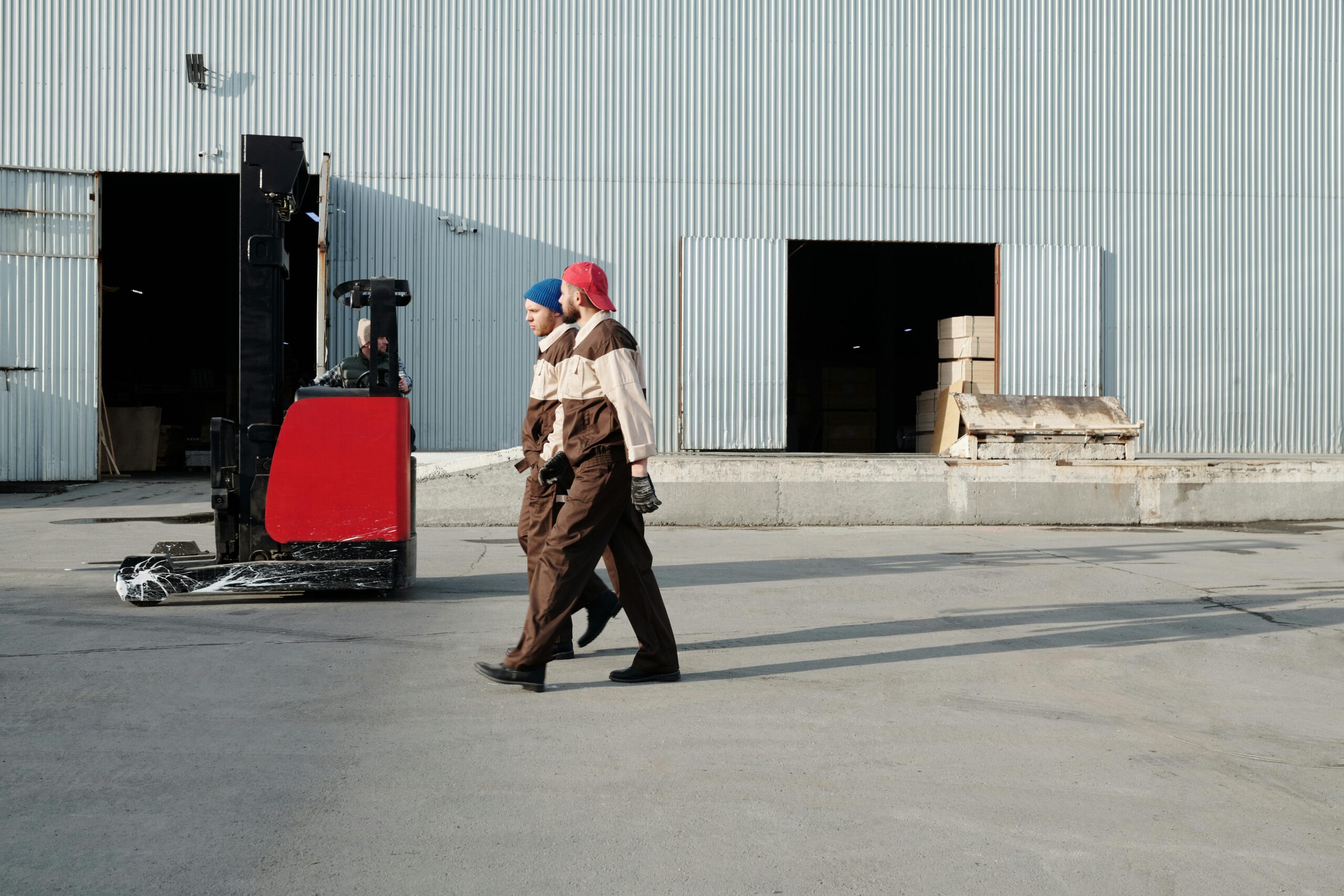 pexels-photo-4487445-4487445 Two workers in uniforms walking outdoors near a forklift at a warehouse.