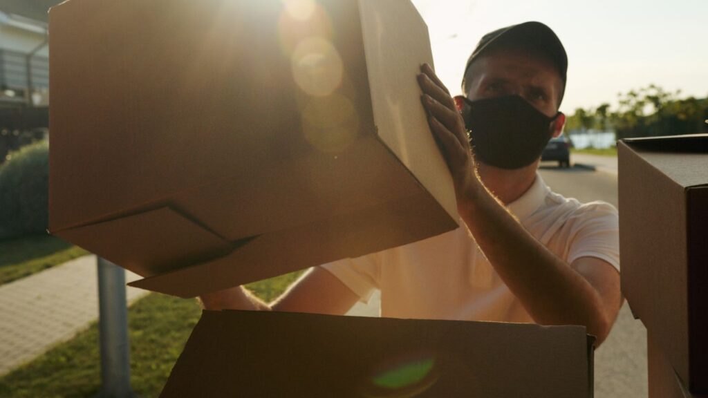 A masked courier delivers packages in bright sunlight, ensuring safe and timely shipping.