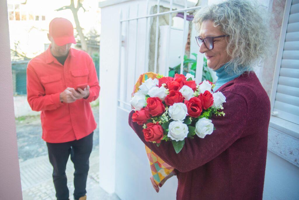 A cheerful elderly woman receives a vibrant bouquet of roses from a courier at her doorstep.