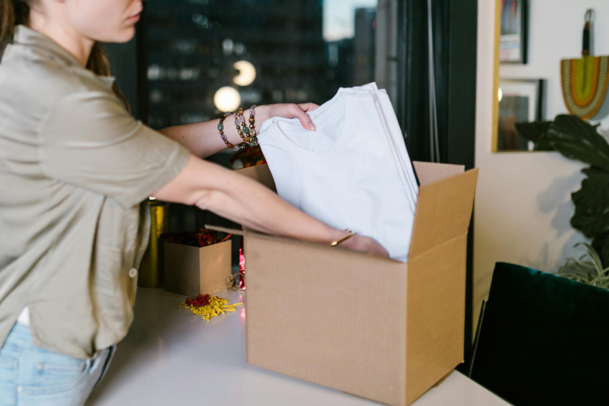 pexels-photo-7310094-7310094 A woman packs white t-shirts into a cardboard box, set in a modern indoor environment.