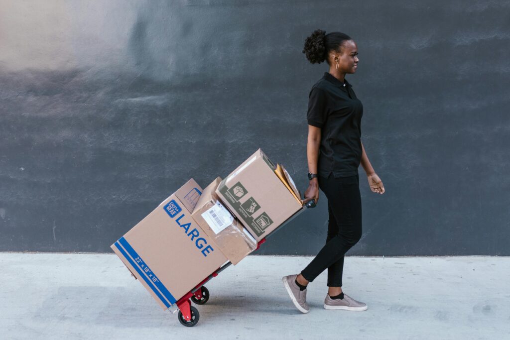 A female courier in uniform sending a large parcel on a trolley outdoors against a dark wall.