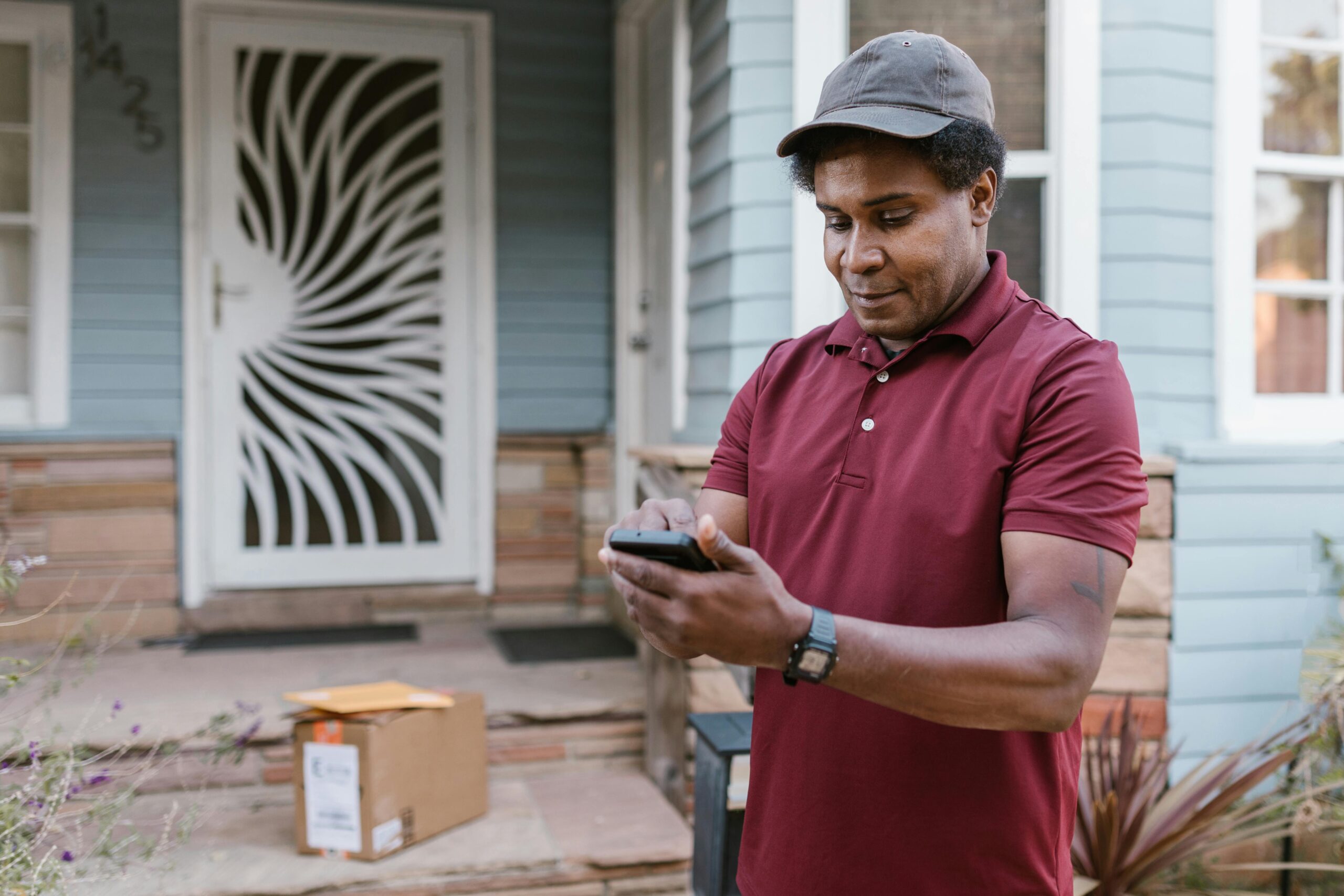 A courier in a maroon shirt checks delivery details on his phone at a residential porch with parcels.