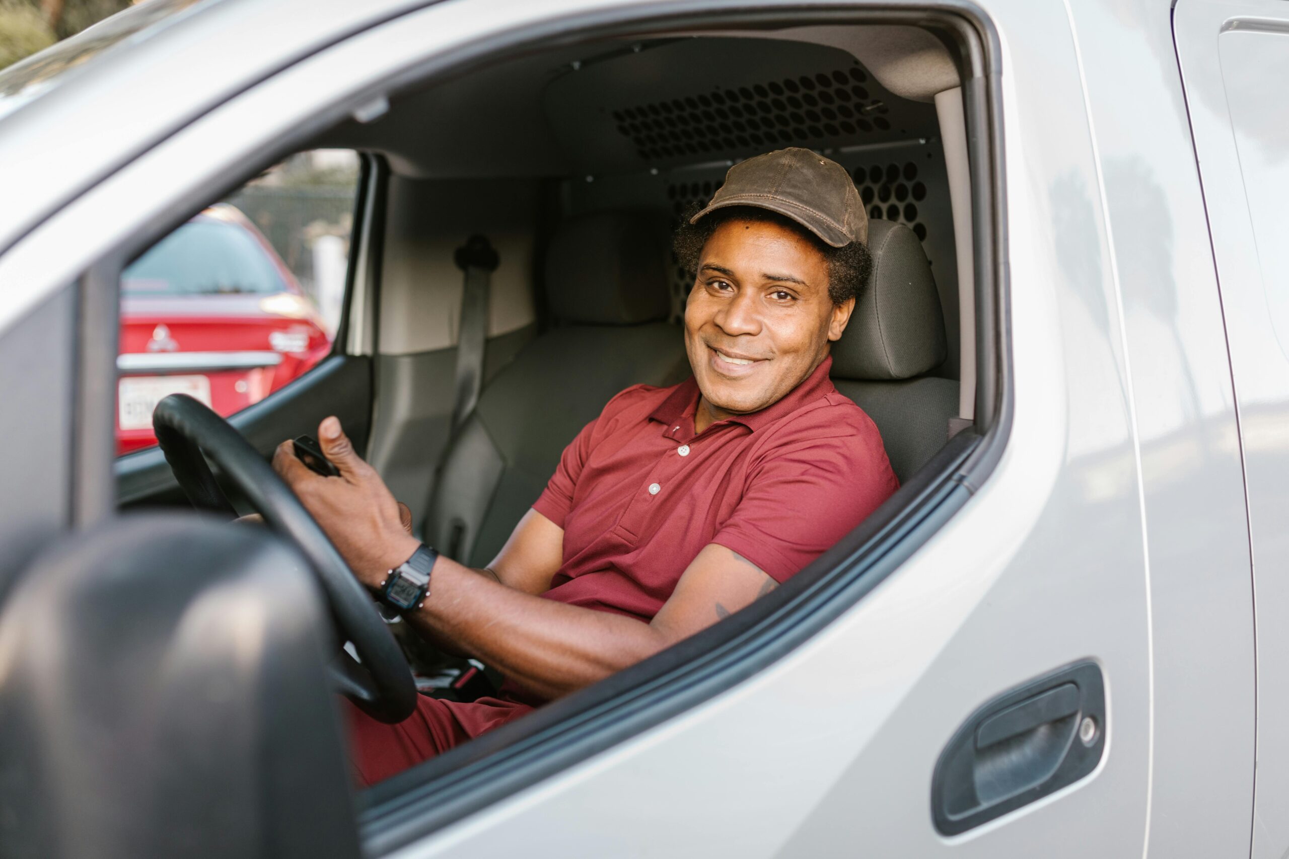 A smiling delivery driver in uniform seated inside his van, holding a smartphone.