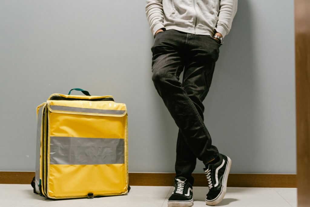Casual food delivery courier leaning against a wall with a yellow thermal bag.