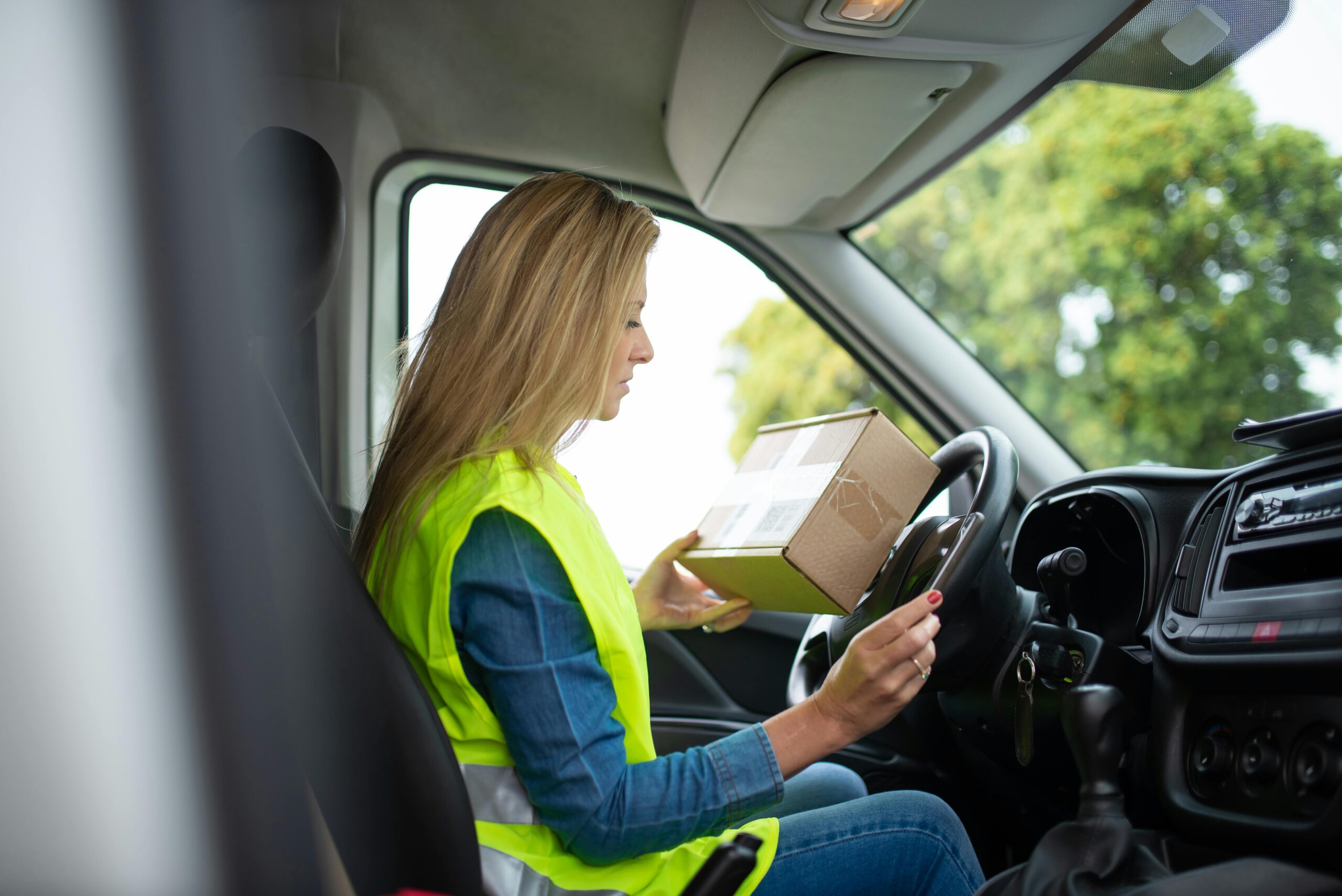 A female delivery driver in Portugal holds a package inside a van, wearing a high-visibility vest.