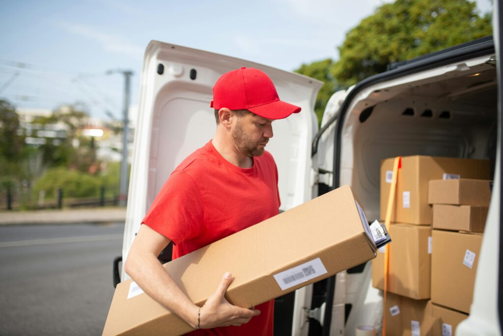 pexels-photo-7843960-7843960-2 Courier in red uniform unloading packages from a delivery van on a sunny day.