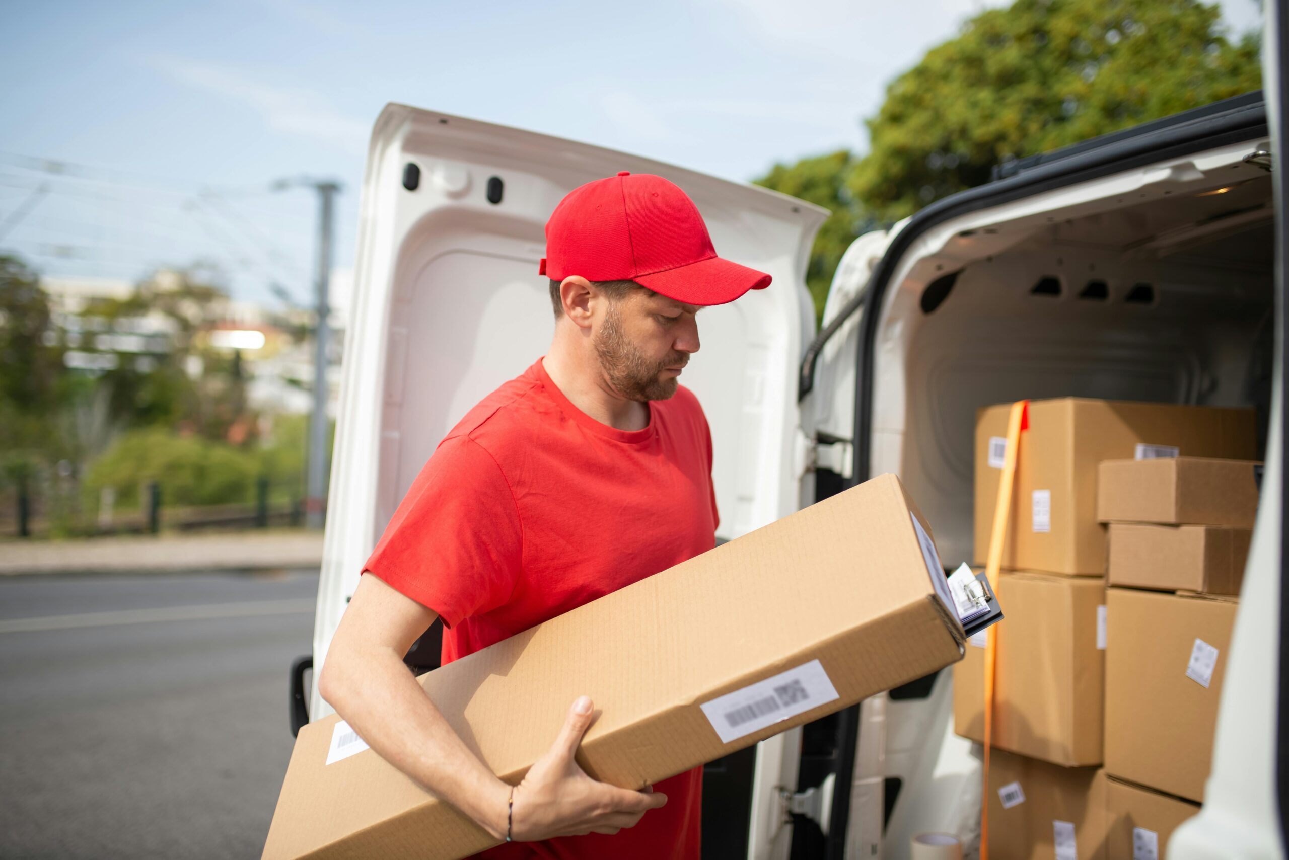 pexels-photo-7843960-7843960 Courier in red uniform unloading packages from a delivery van on a sunny day.