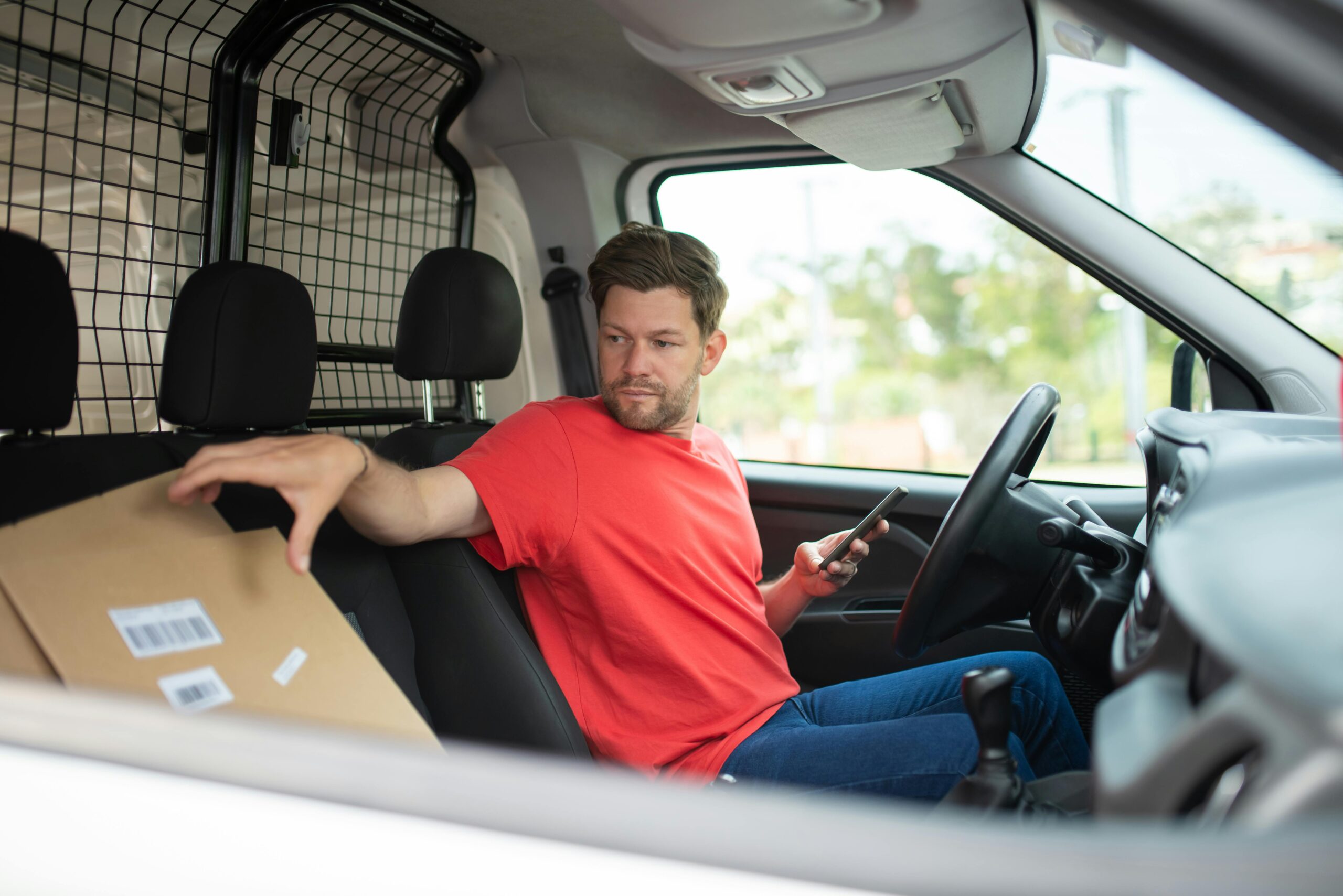 pexels-photo-7843963-7843963 Man in red shirt handling package inside delivery van, holding phone.