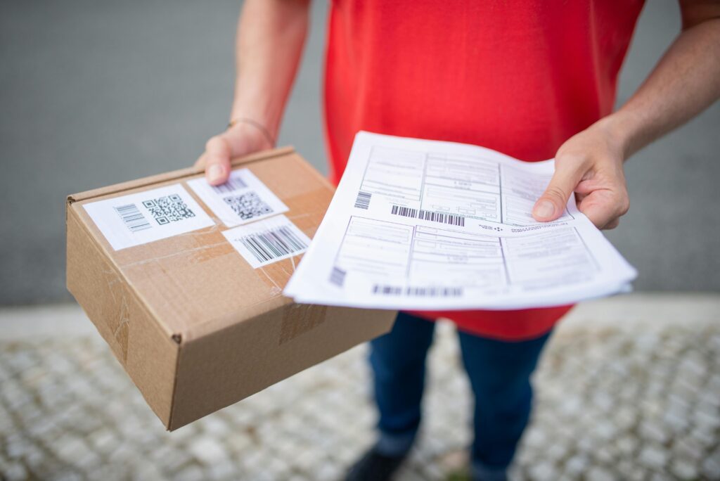 pexels-photo-7843991-7843991 Close-up of person in red shirt holding a brown package and delivery documents outside.