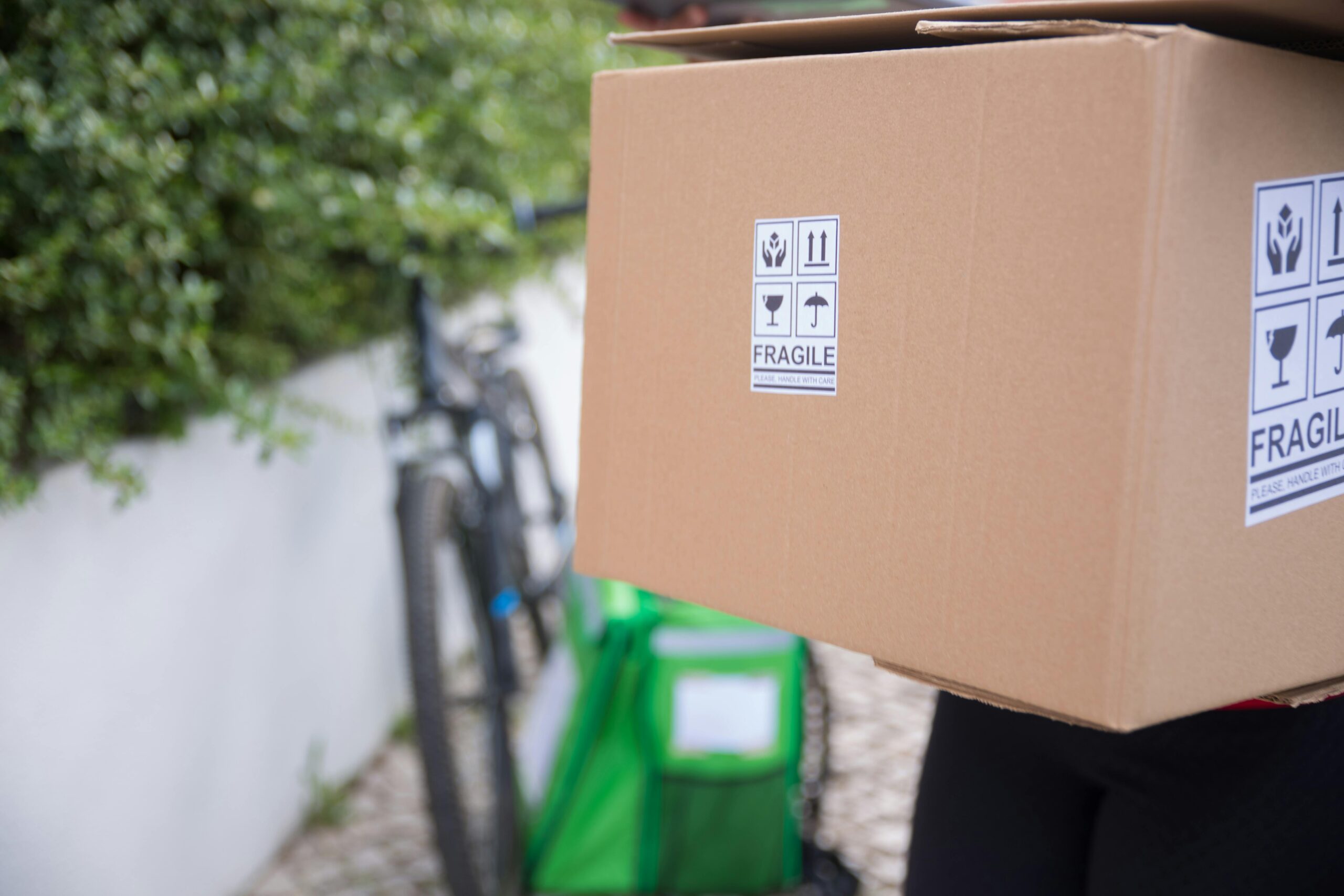 A courier delivers a fragile cardboard box with a bicycle parked beside on a sunny day.